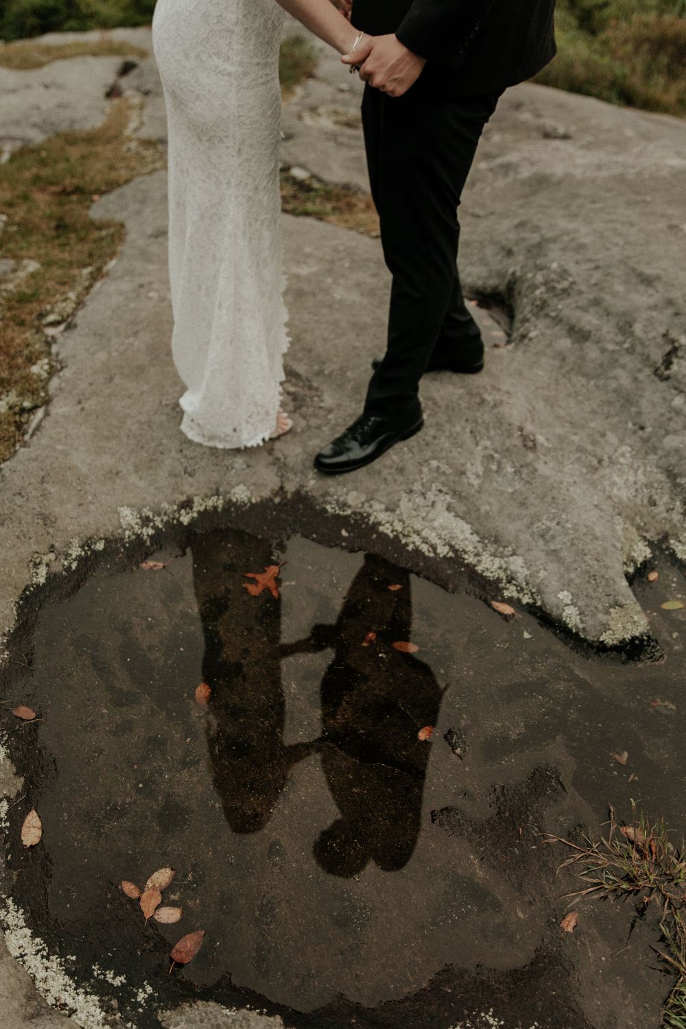 Reflections of a bride and groom's feet in a puddle surrounded by autumn leaves on stone ground.