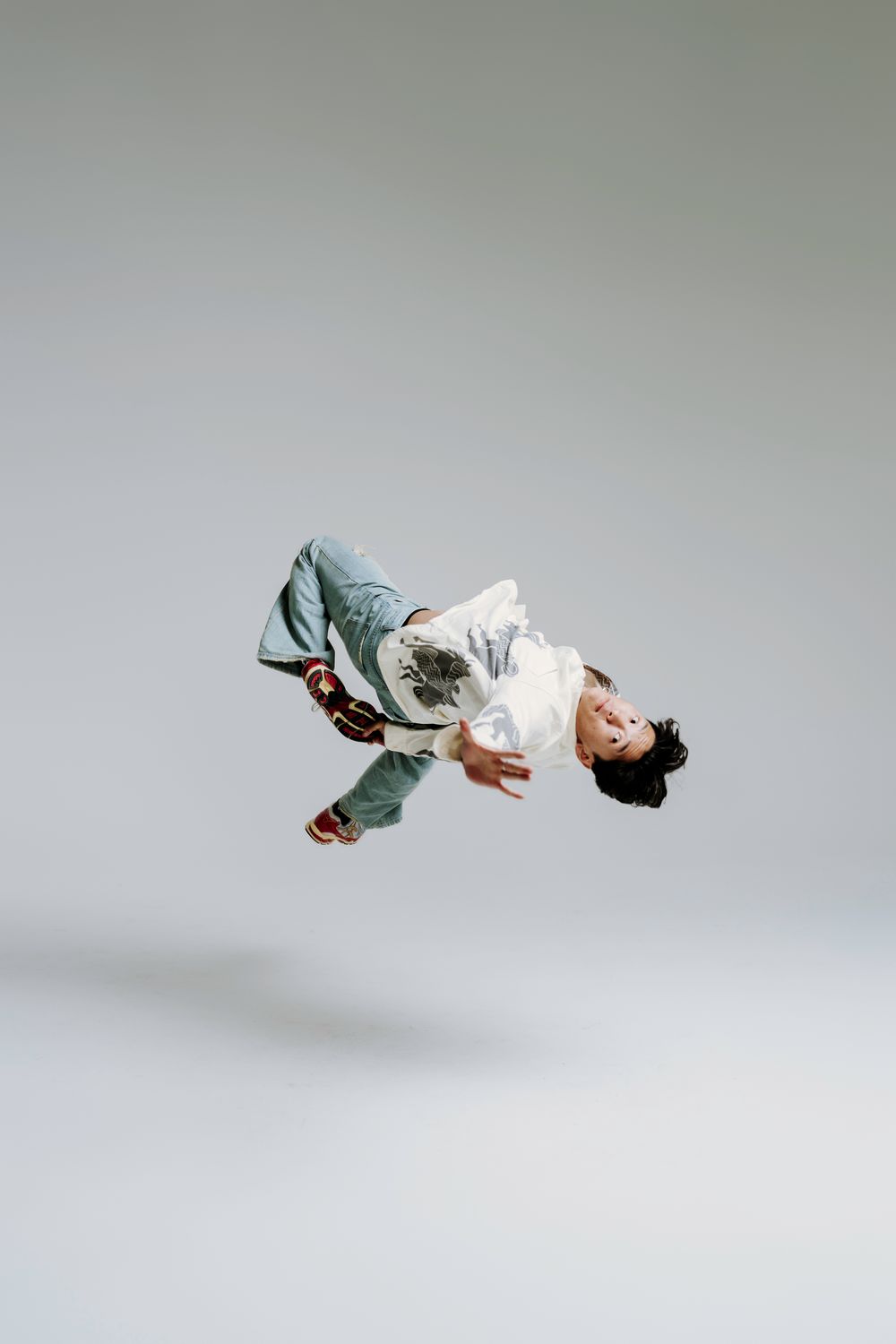 Person in loose blue clothing performing an artistic aerial backflip against a light gray studio background.