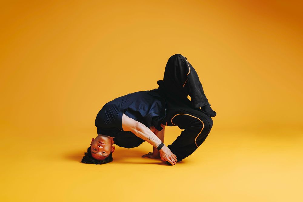 Dancer in black hoodie and pants performs break dance move on vibrant yellow background.