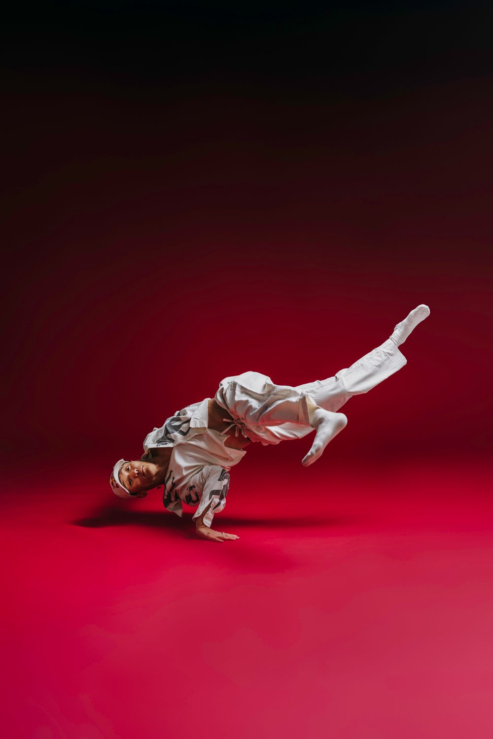 A person in white martial arts uniform performs a dynamic breakdancing move against a dark red background.