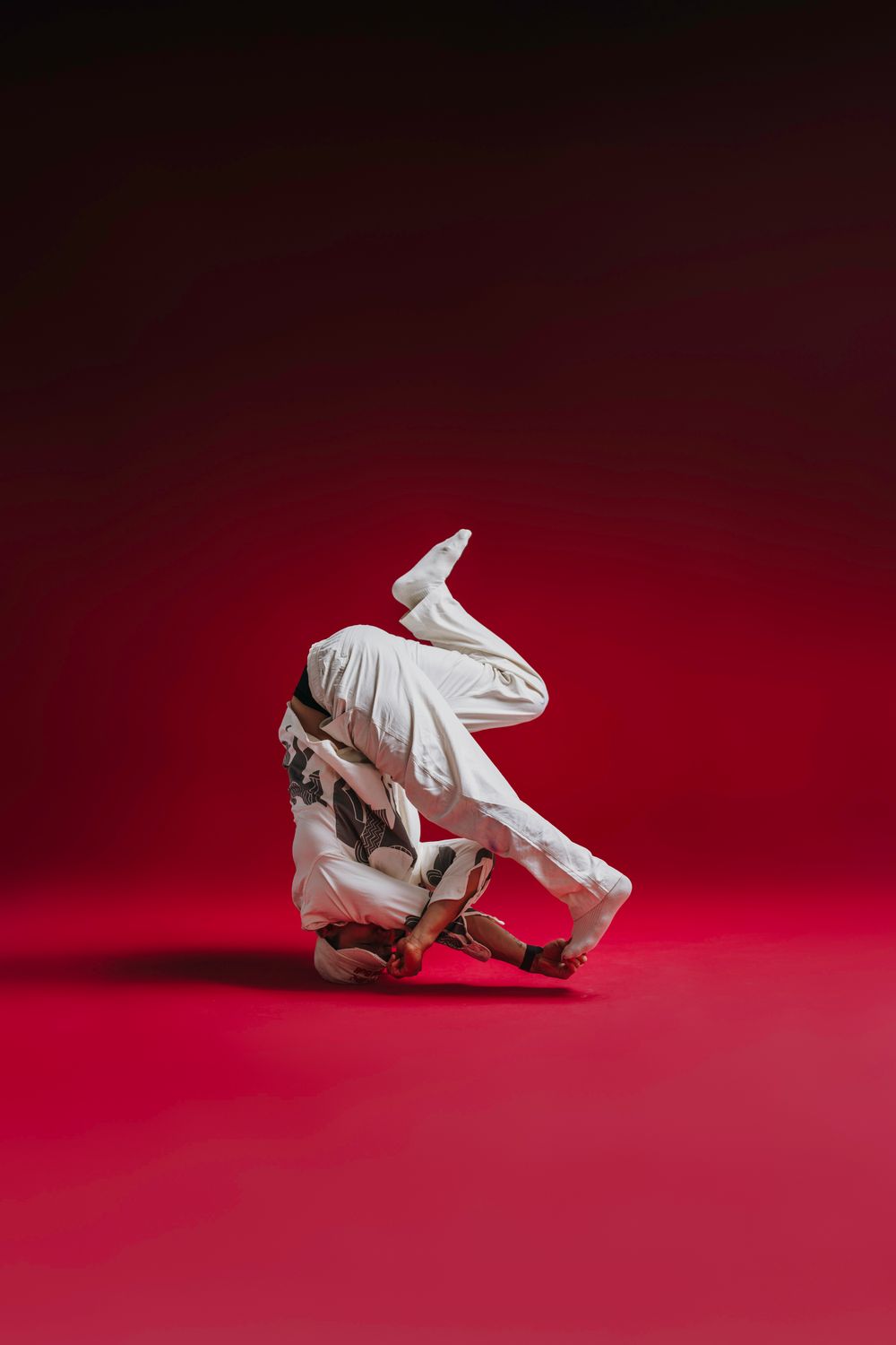 Person in white clothing performs an athletic breakdancing freeze move against a dramatic red background.