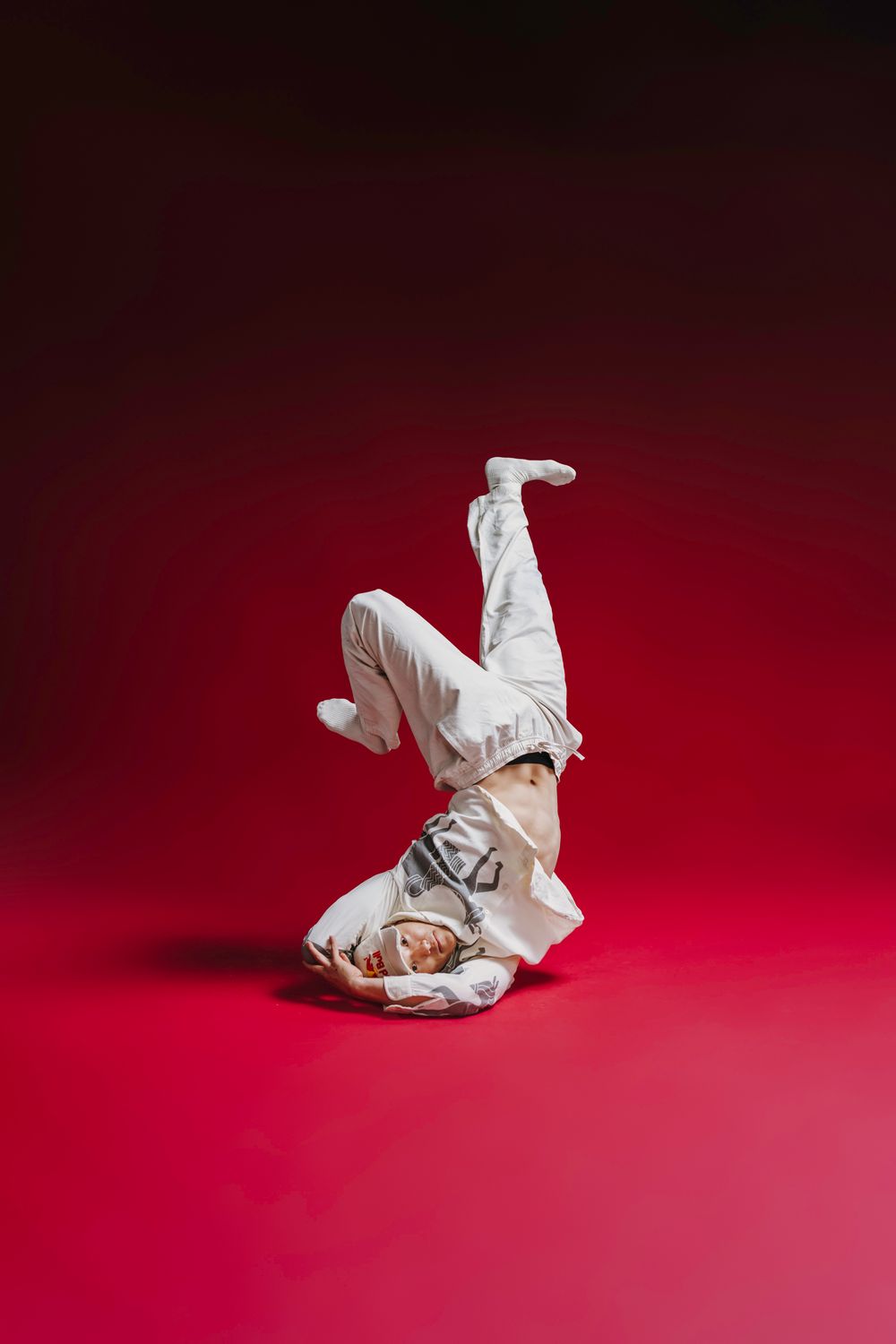 Dancer in white outfit performing a dynamic breakdance freeze move on a vibrant red background.