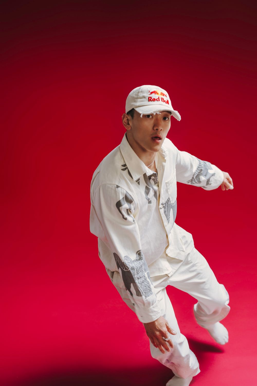 Dancer in white outfit and cap performs energetic move against vibrant red background.