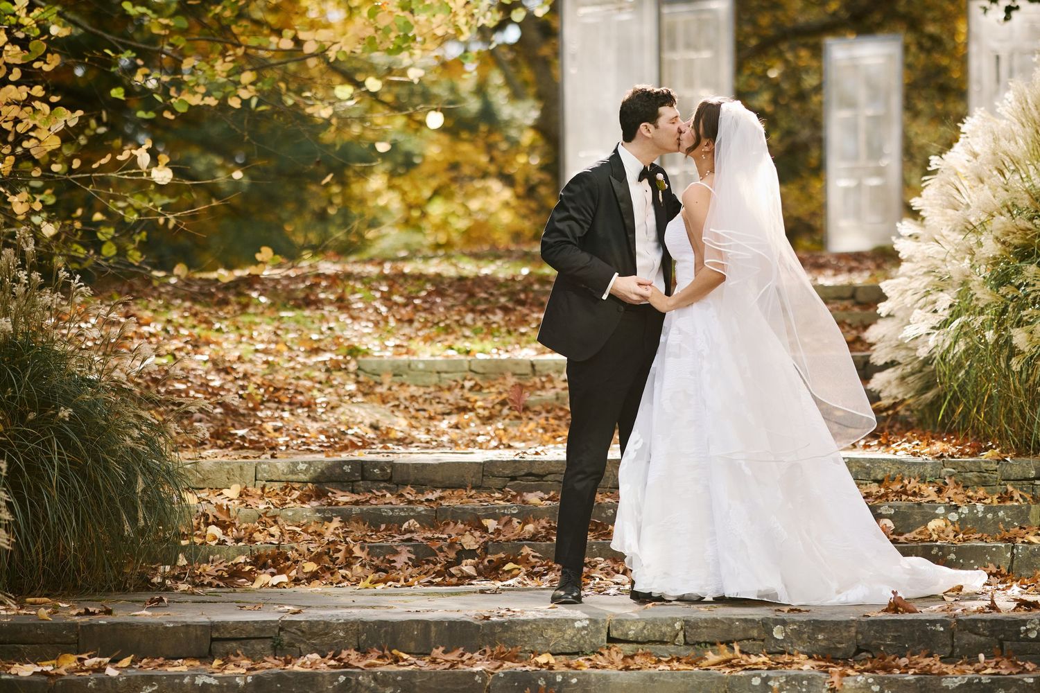 A couple in wedding attire share an intimate moment in a garden setting with golden autumn foliage.