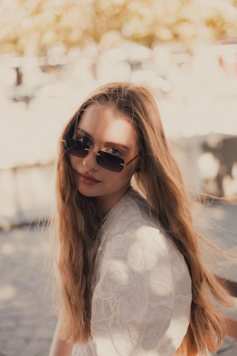 Stylish woman in white dress and sunglasses with long hair in warm sunlight.