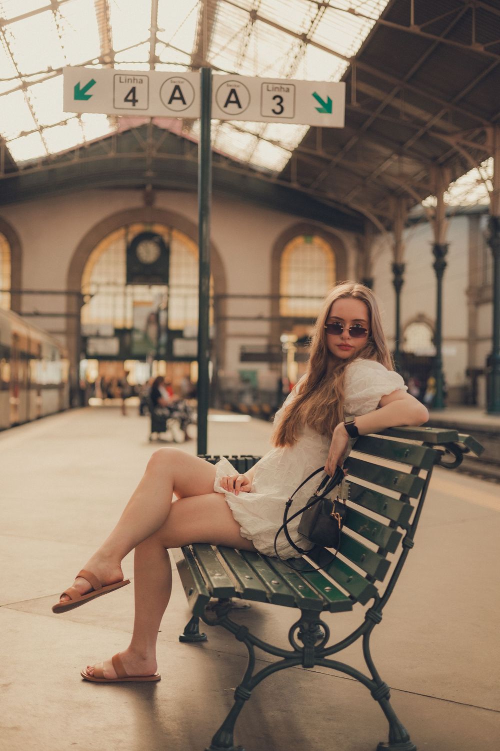 Woman in white dress sitting on vintage train station bench under directional signs.