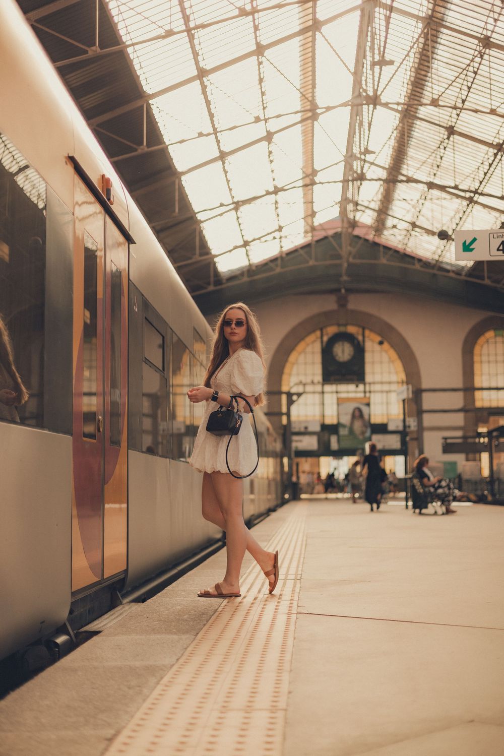 Woman in white dress standing on train platform with architectural glass ceiling.