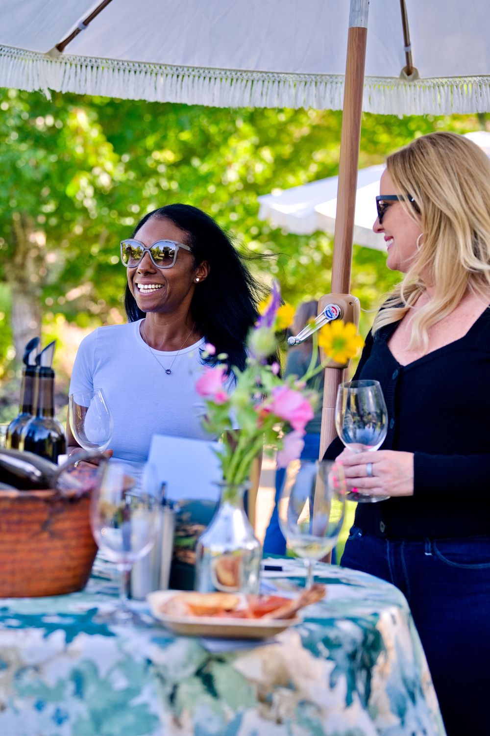 Two friends share a laugh while enjoying wine and appetizers under a white umbrella at an outdoor table with flowers.