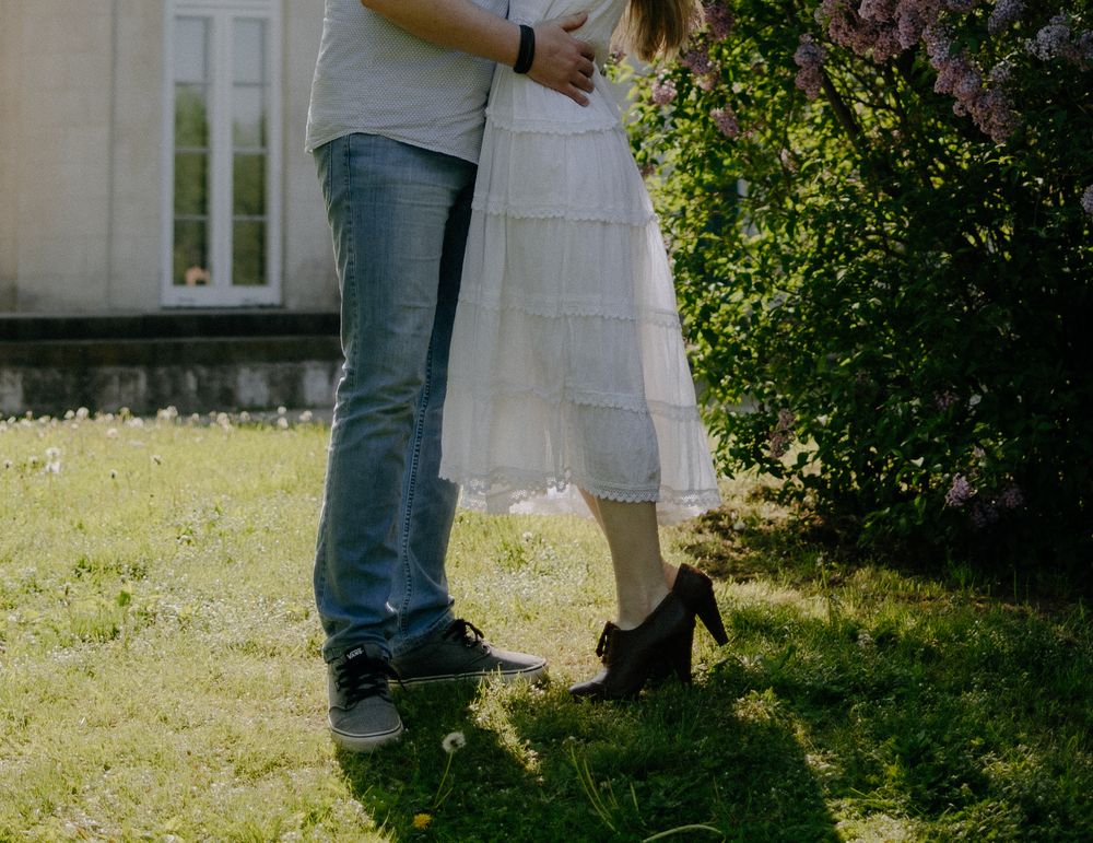 Two people standing close together on grass, one wearing blue jeans and the other in a flowing white skirt and heels.