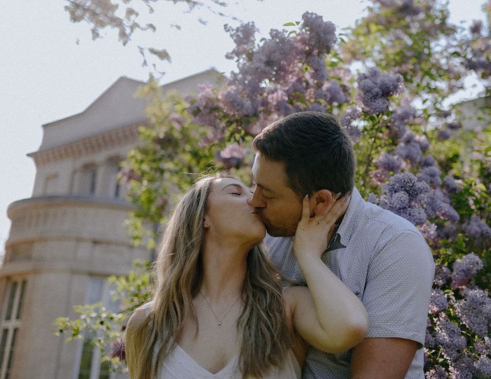 A romantic couple shares a tender kiss among blooming purple wisteria flowers with a classical building in the background.