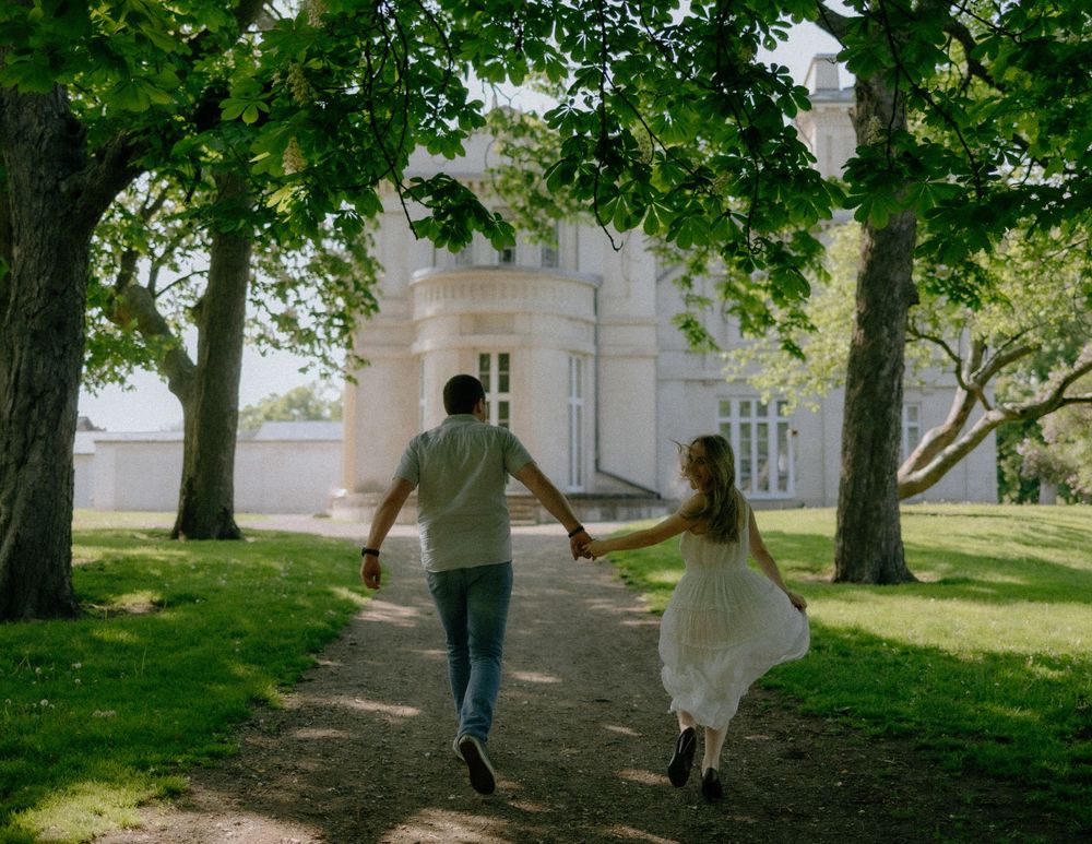 A couple runs playfully hand in hand down a tree-lined path towards a grand white mansion on a sunny day.