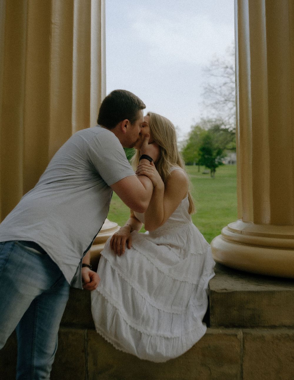 Couple sharing an intimate moment on a stone pillar against a green landscape backdrop at dusk.