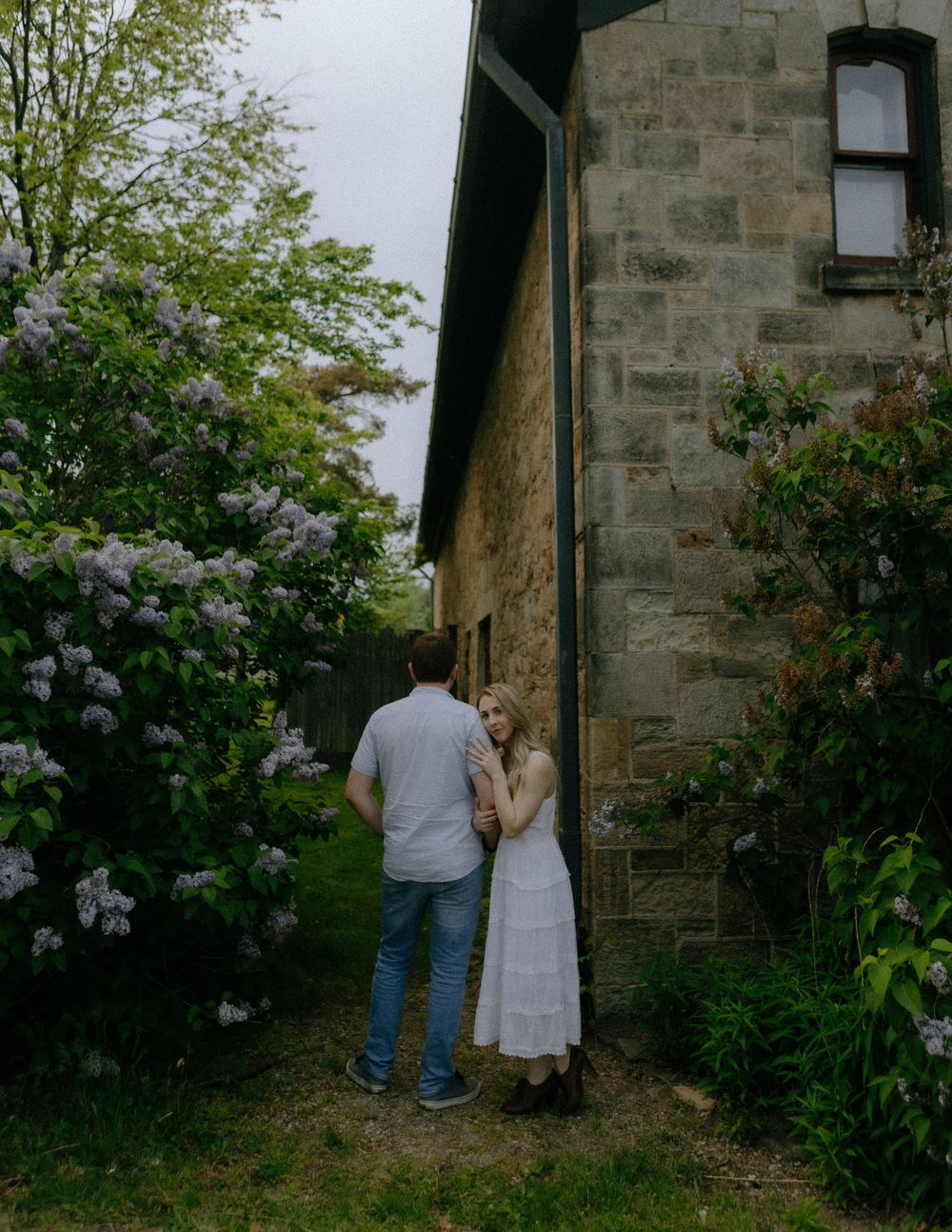 A couple stands in front of a stone house with blooming lilac bushes in a garden setting.
