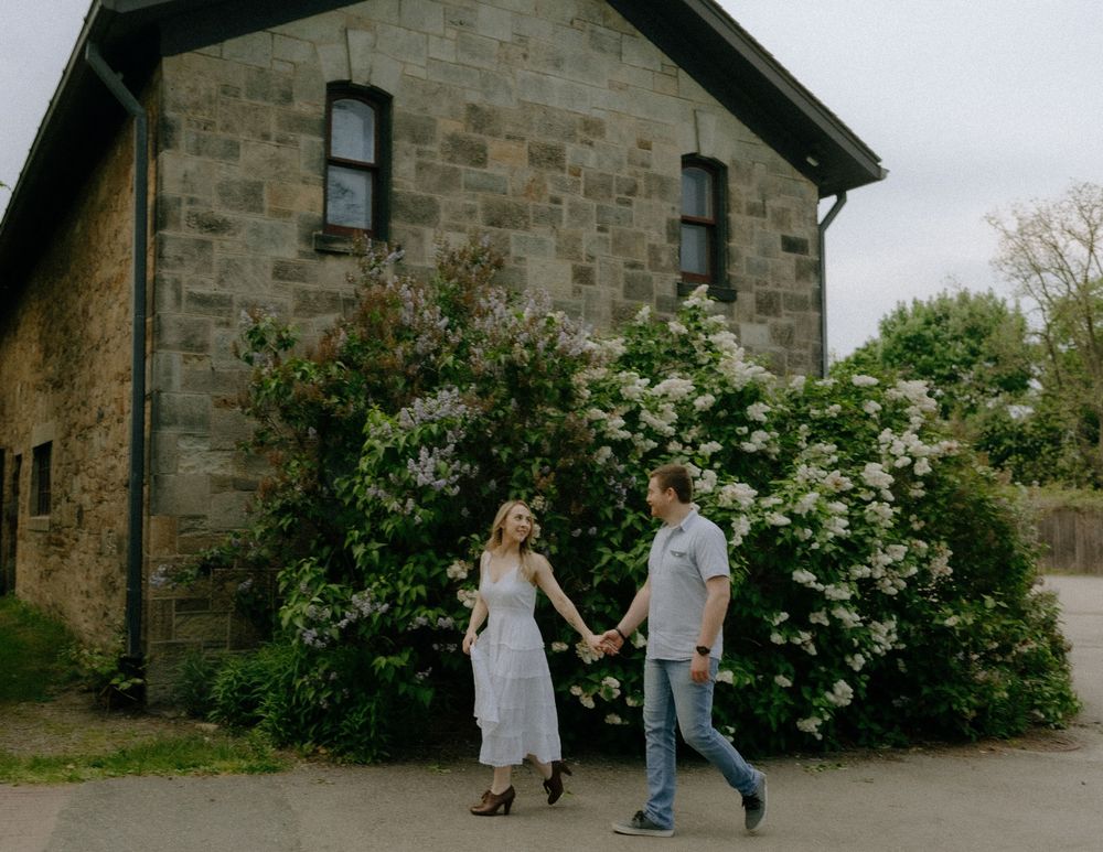 A couple playfully runs and embraces in front of a rustic stone building with blooming bushes.