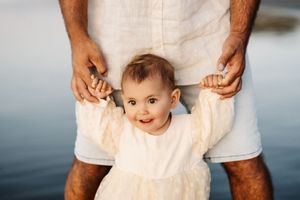 A baby in white dress reaches up with joy while supported by caring hands.