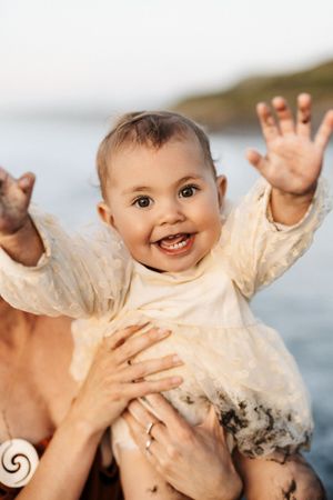 A happy baby in a lace dress reaches up with excitement against a coastal background.