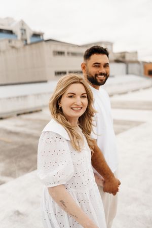 A couple wearing white outfits smile while standing on a rooftop with urban buildings in background.
