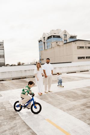 Family watches child riding blue bicycle on urban rooftop with city buildings in background.