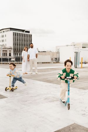 Children riding scooters on Hamilton rooftop carpark while adults watch in background.