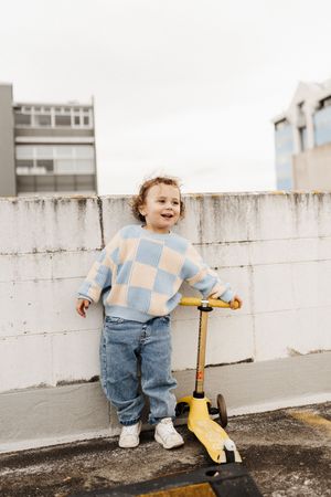 Toddler in checkered sweater smiling while holding yellow scooter on rooftop.
