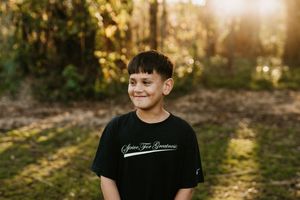 Young person in black t-shirt smiling outdoors with sunlit trees in background.