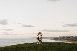 Romantic couple standing on grassy cliff overlooking ocean at sunset.