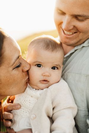 A baby in a white sweater being lovingly embraced by parents in warm sunset lighting.