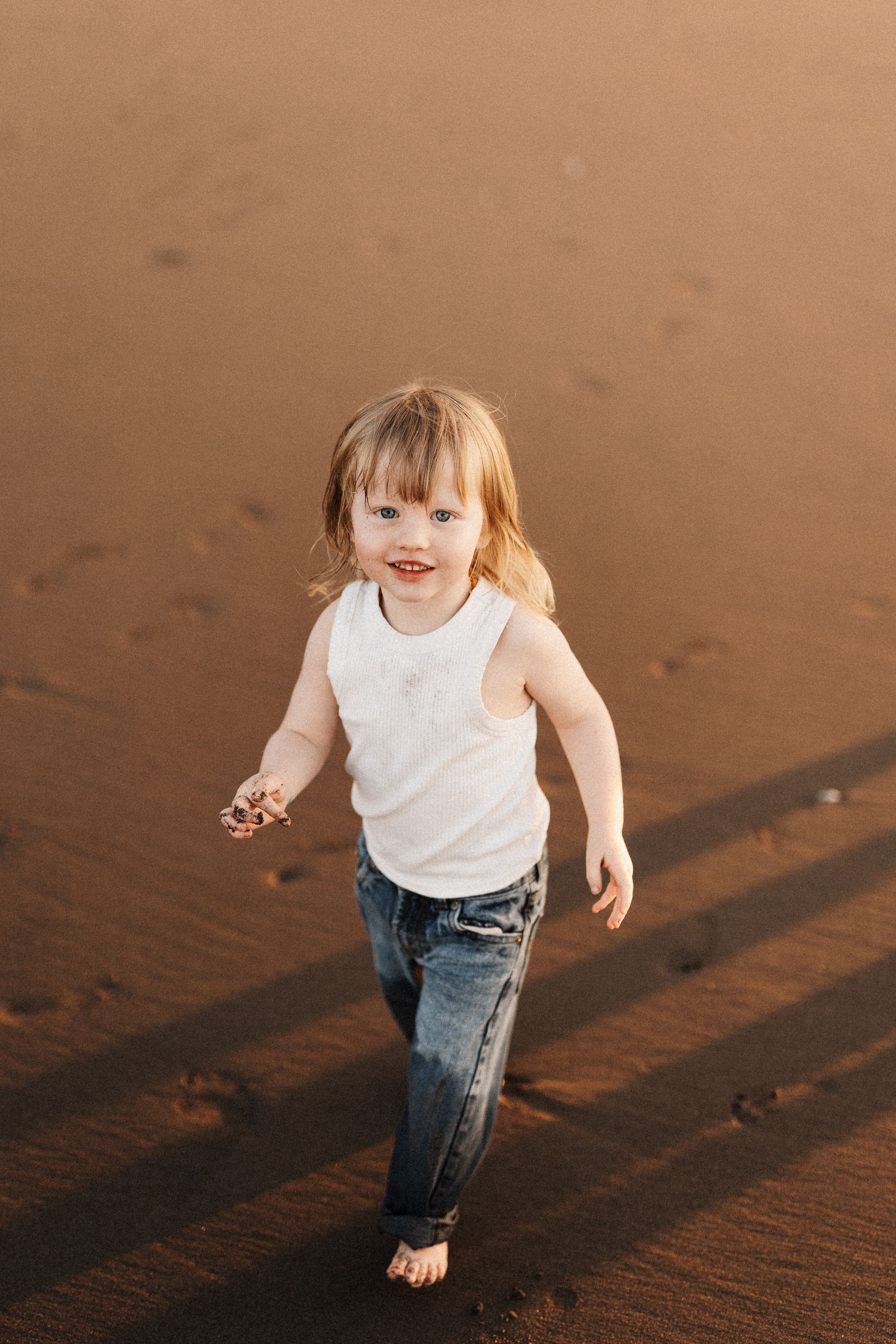 Small child in white tank top and jeans running joyfully on sand.