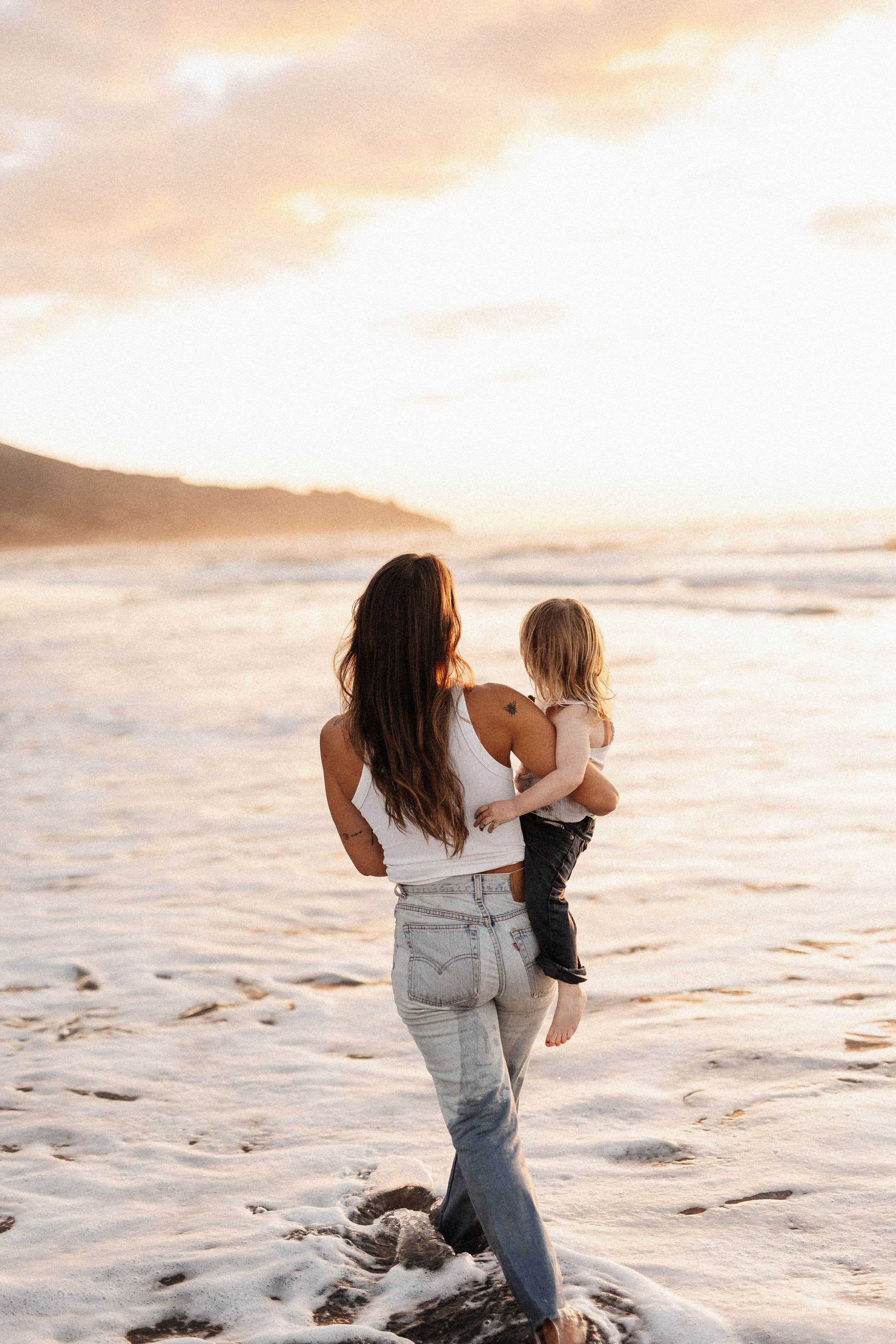 Mother carrying child while standing in ocean waves during Raglan sunset.