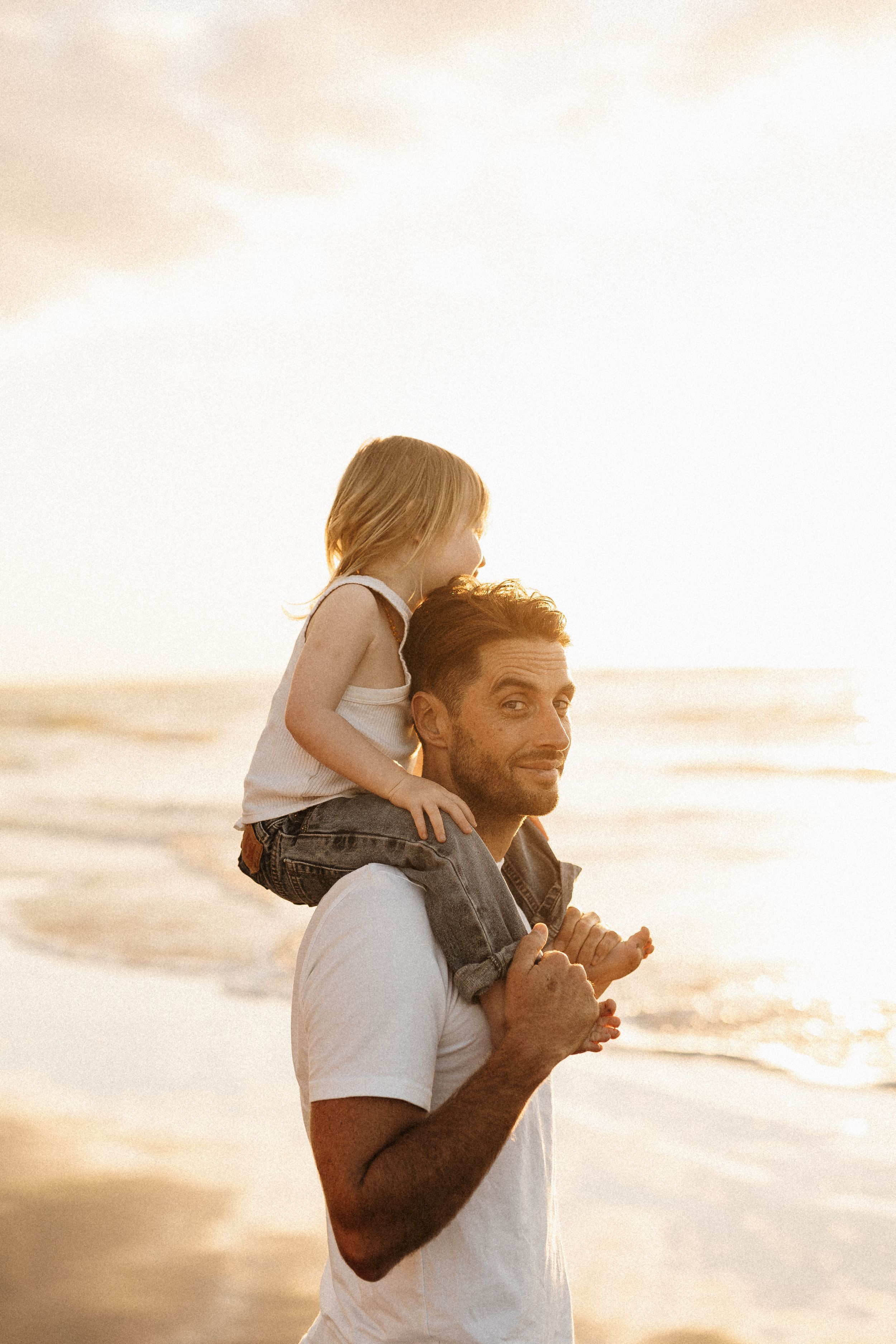A toddler rides on shoulders during a golden sunset at the beach.