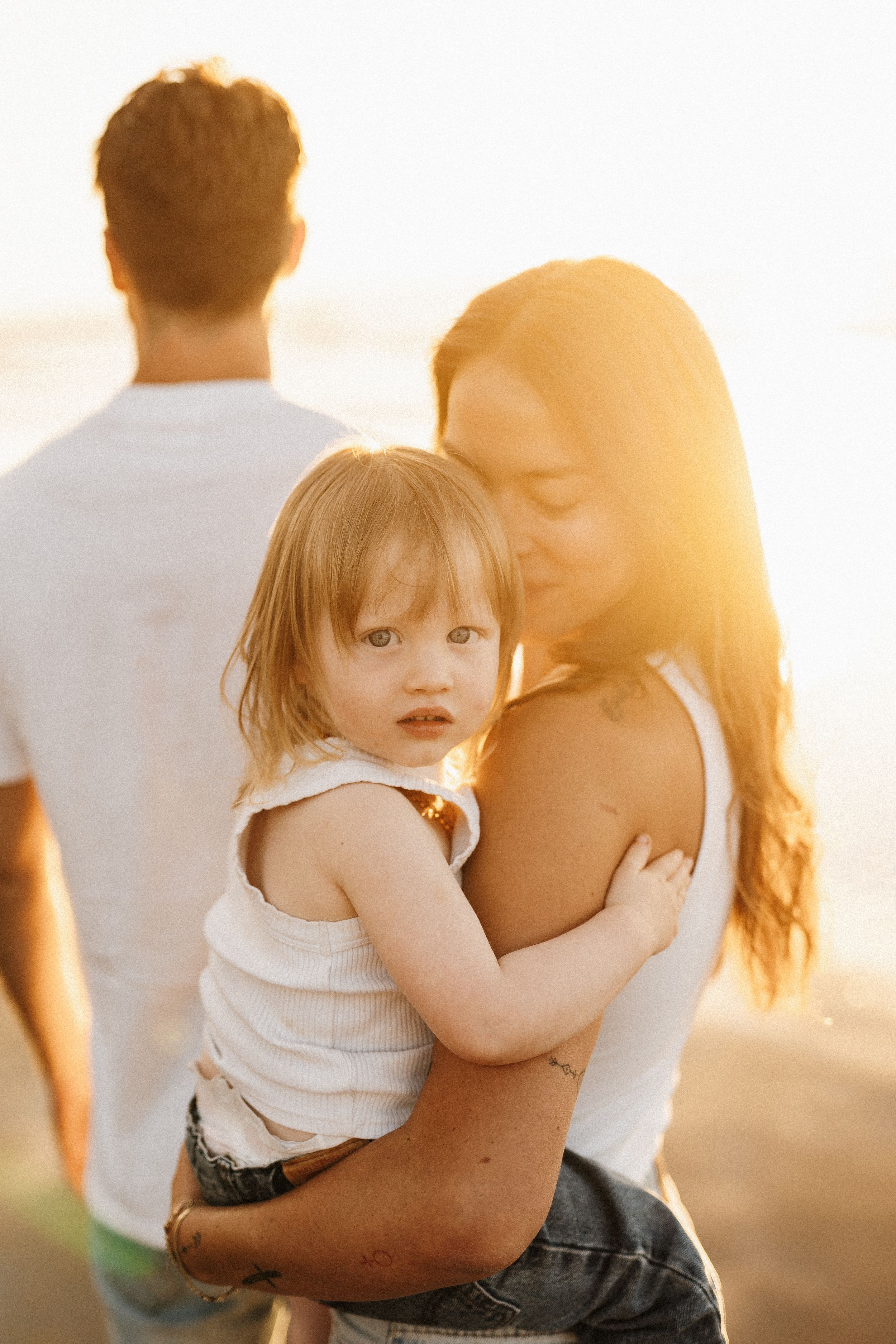 Mother holding toddler at beach during golden hour as father stands nearby.