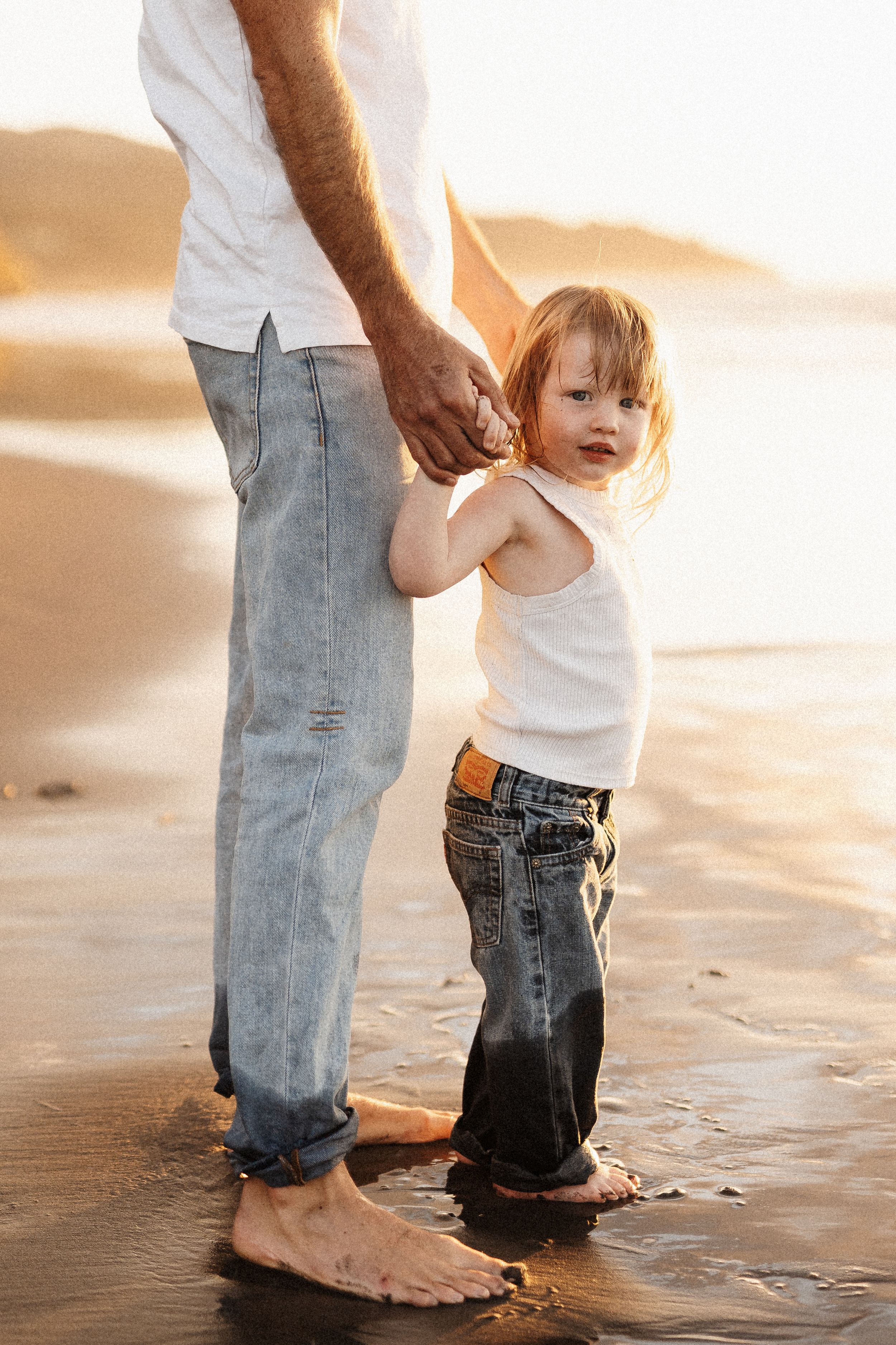 Adult and child holding hands while standing barefoot in wet sand at the beach.