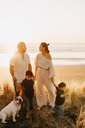 Family and their dog gather among coastal grass at Raglan sunset by the ocean.