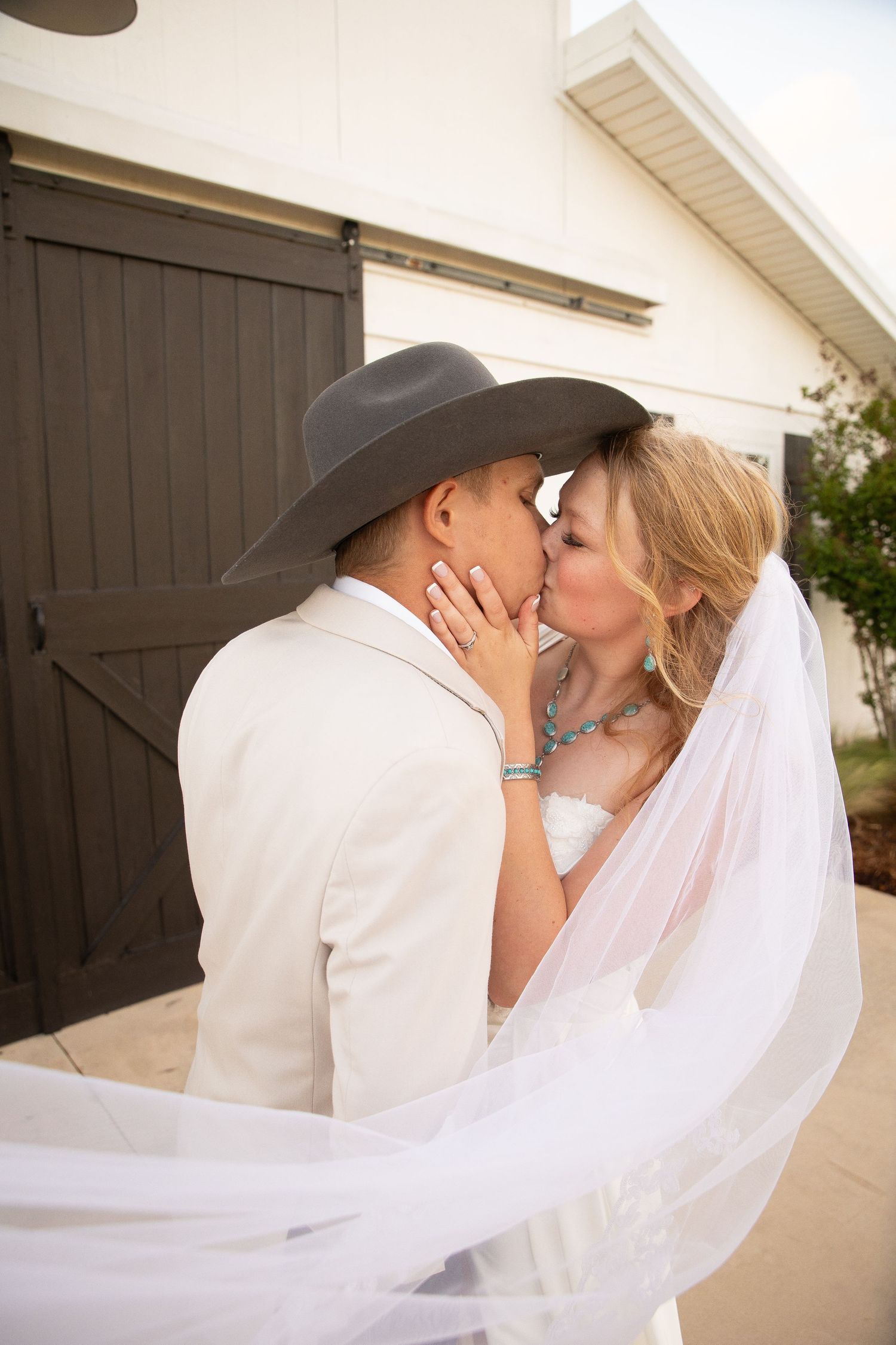 A romantic wedding couple in western attire share tender kisses under a white flowing veil against a light building exterior.