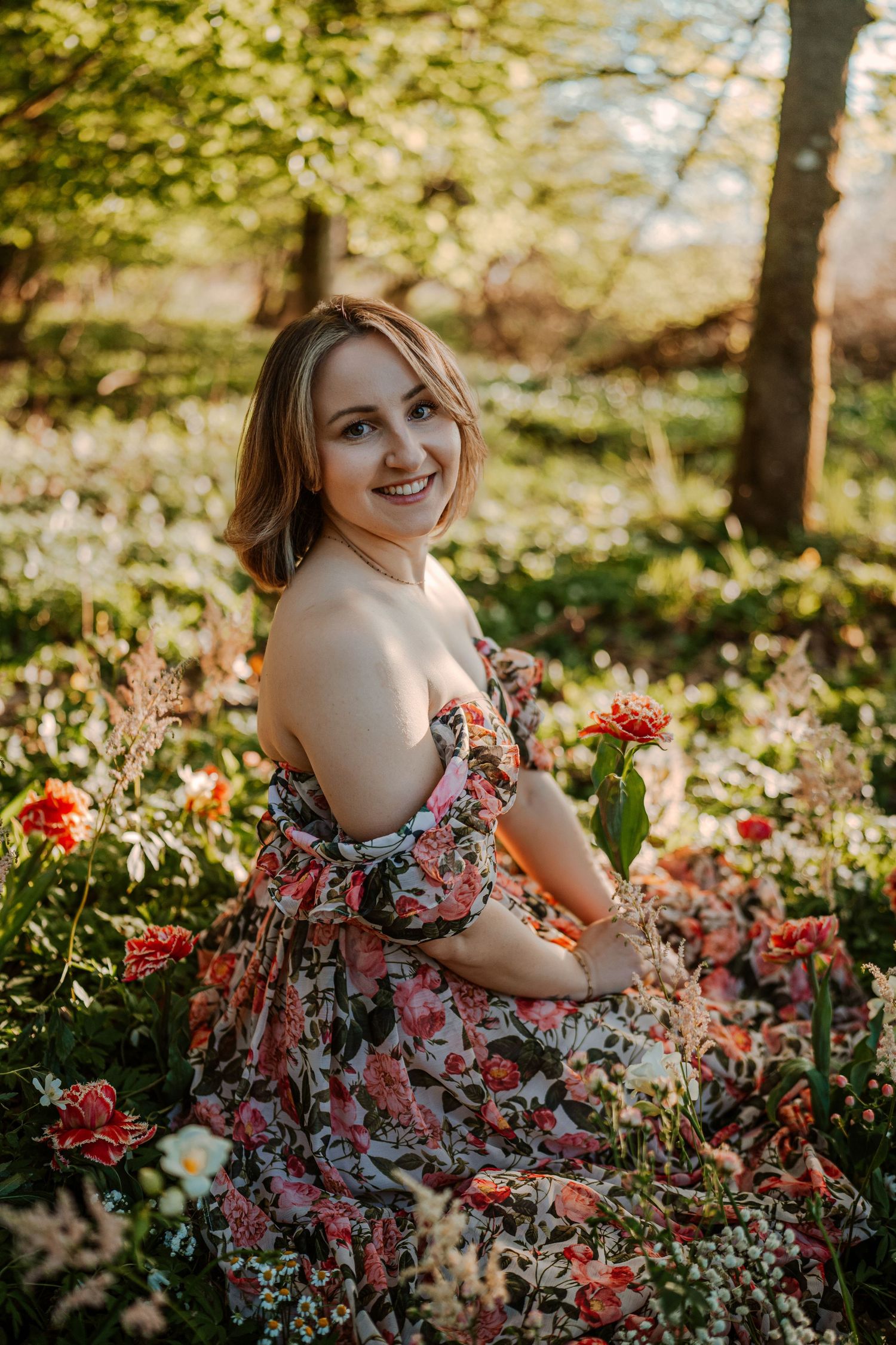 A person in a floral off-shoulder dress poses in a blooming garden during golden hour among pink and red flowers.