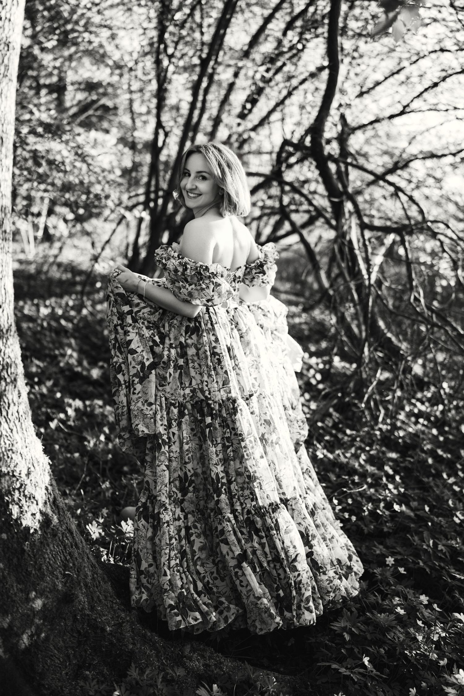 A person in a flowing lace dress stands in a dreamy forest setting captured in black and white photography.