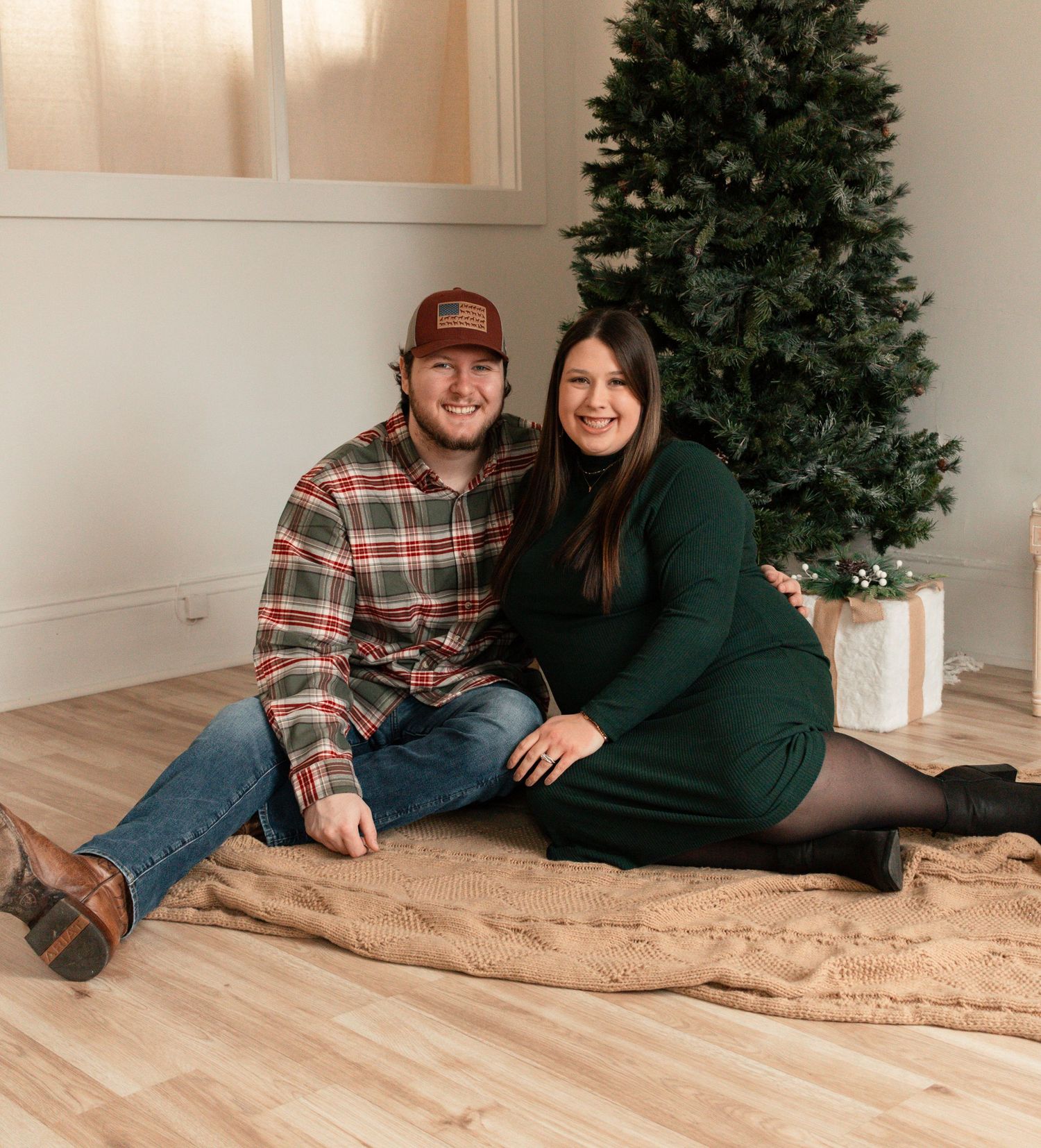 A couple in casual winter attire lounges together on a brown blanket near a Christmas tree in a cozy room.