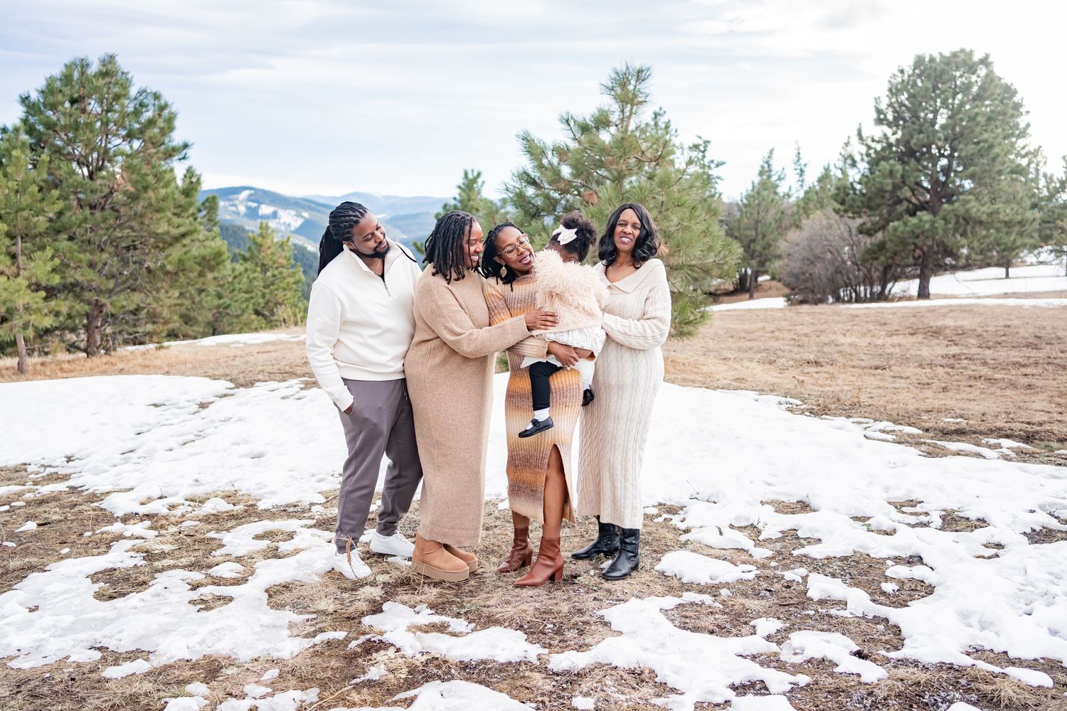A group in coordinating neutral outfits stands together laughing while posing outdoors on snowy ground with pine trees behind them.