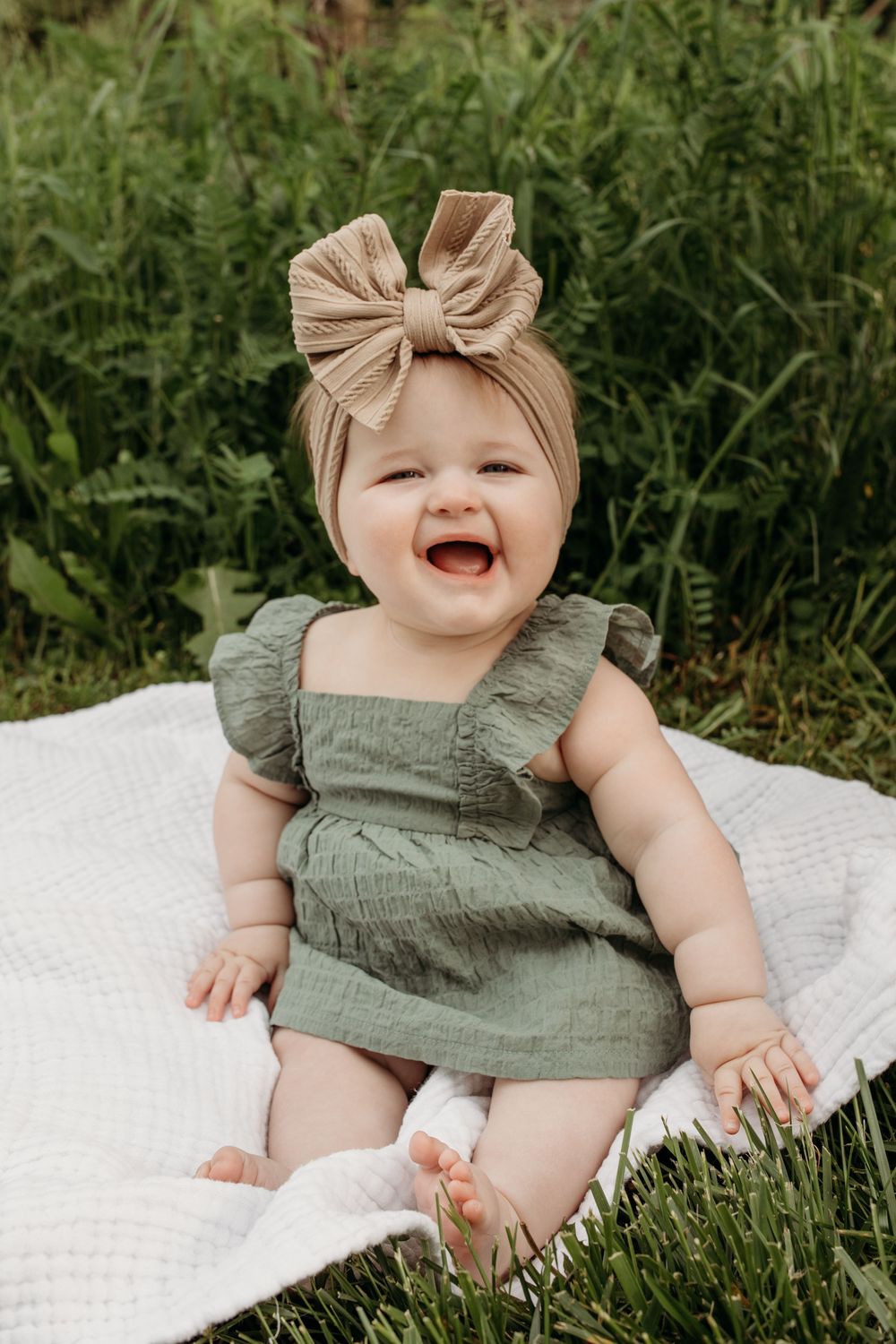 Baby wearing sage green outfit and tan bow laughing while laying on white blanket outdoors.