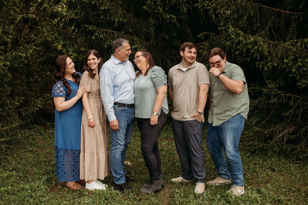 A casual family group shares laughter together while standing in front of green foliage outdoors.