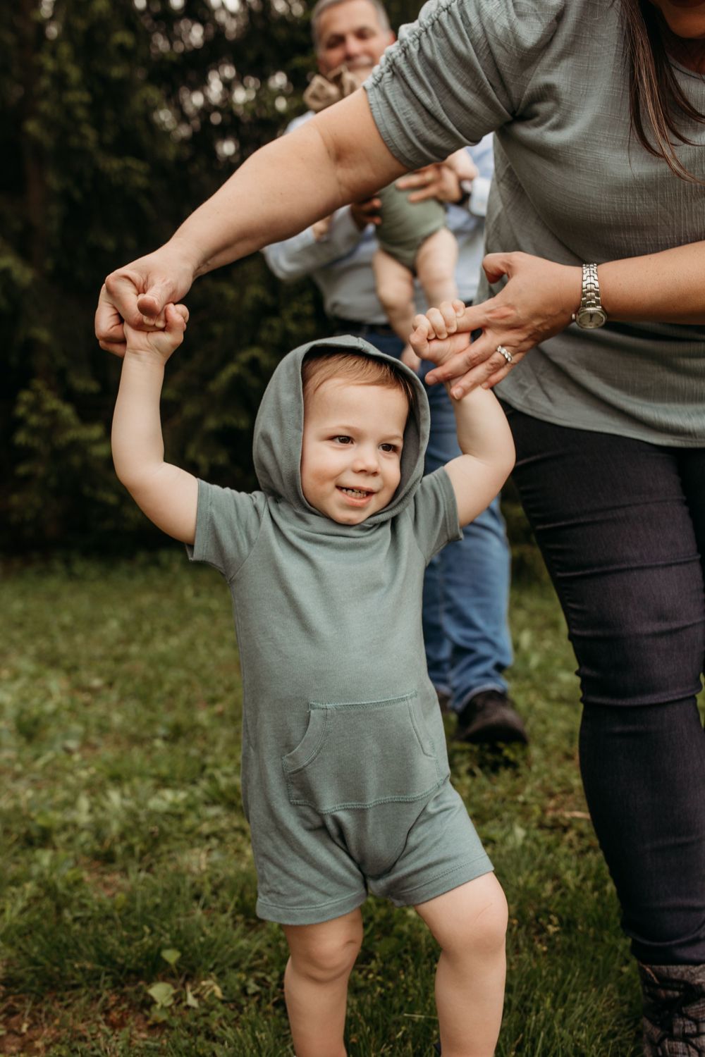 A toddler in a gray hooded romper smiles while holding hands with adults outdoors.