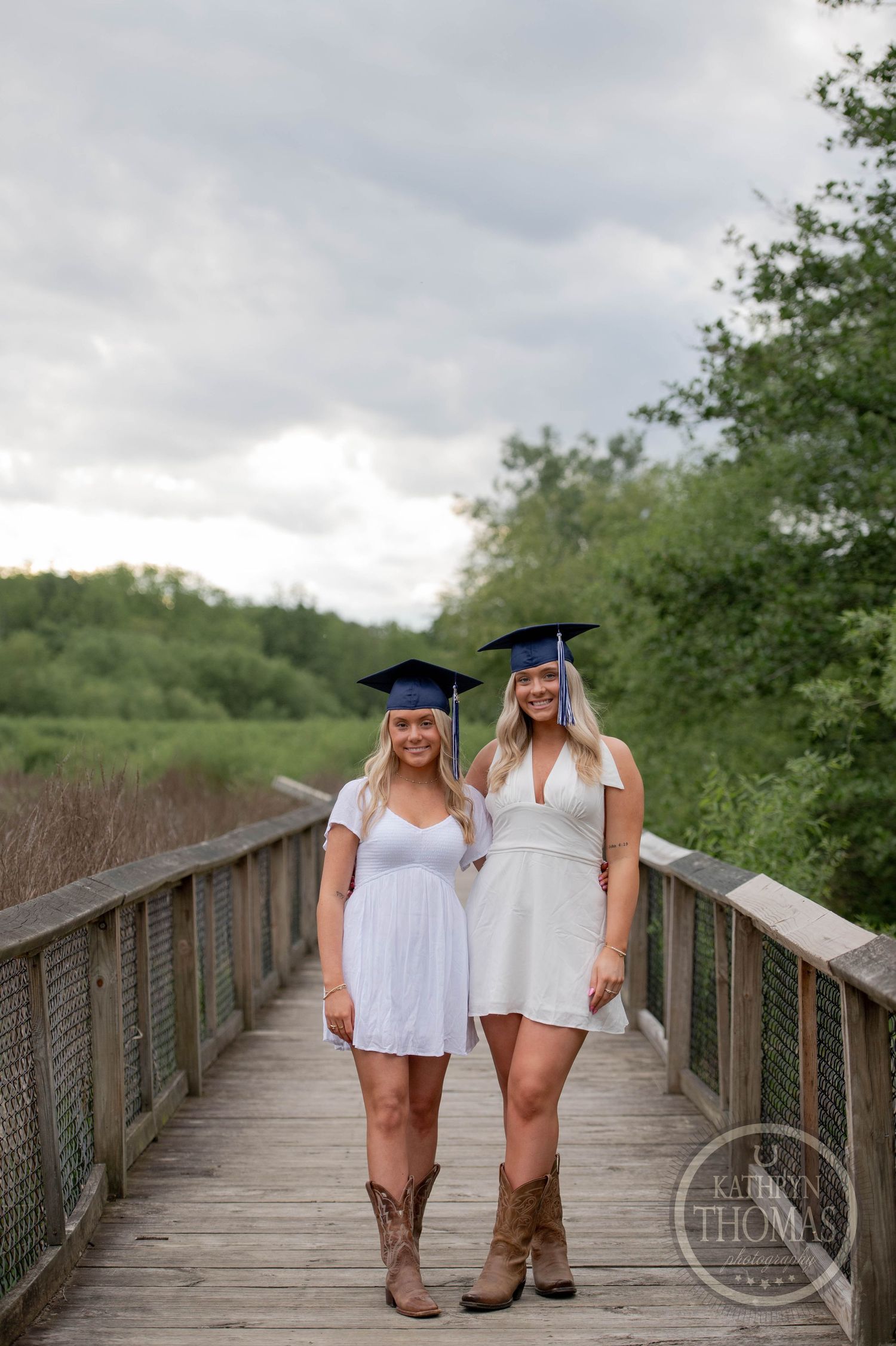 Two graduates wearing white dresses and cowboy boots pose on a wooden boardwalk surrounded by greenery.