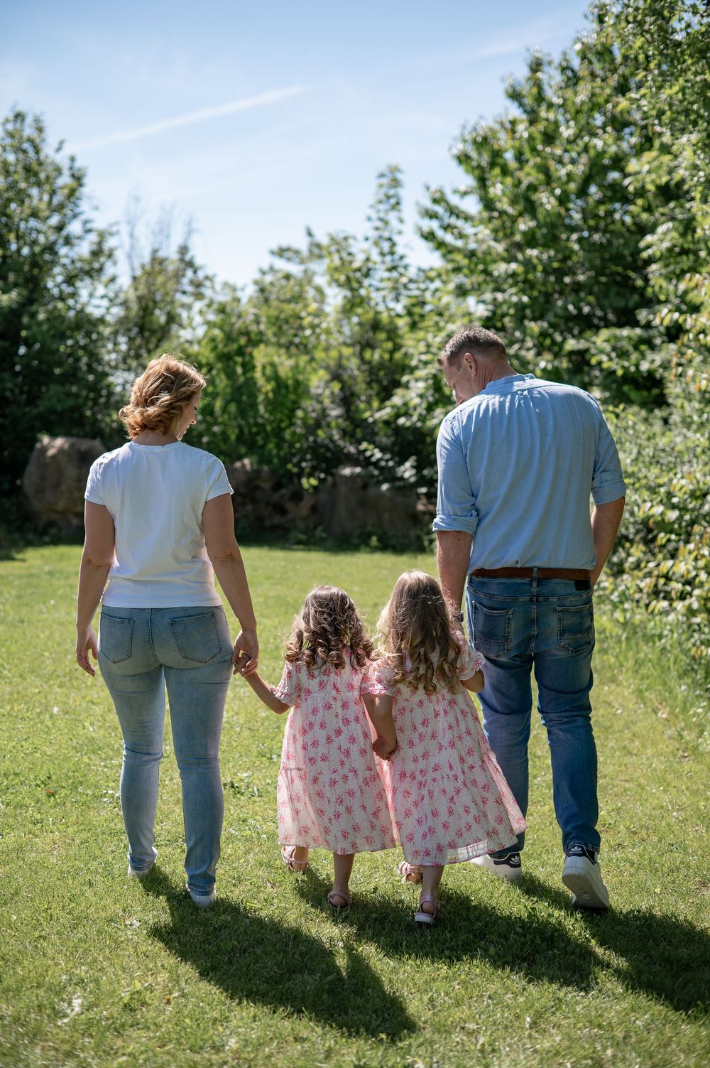 Familie, die an einem sonnigen Tag in einem üppigen grünen Garten spazieren geht, von hinten gesehen, während sie sich an den Händen halten.