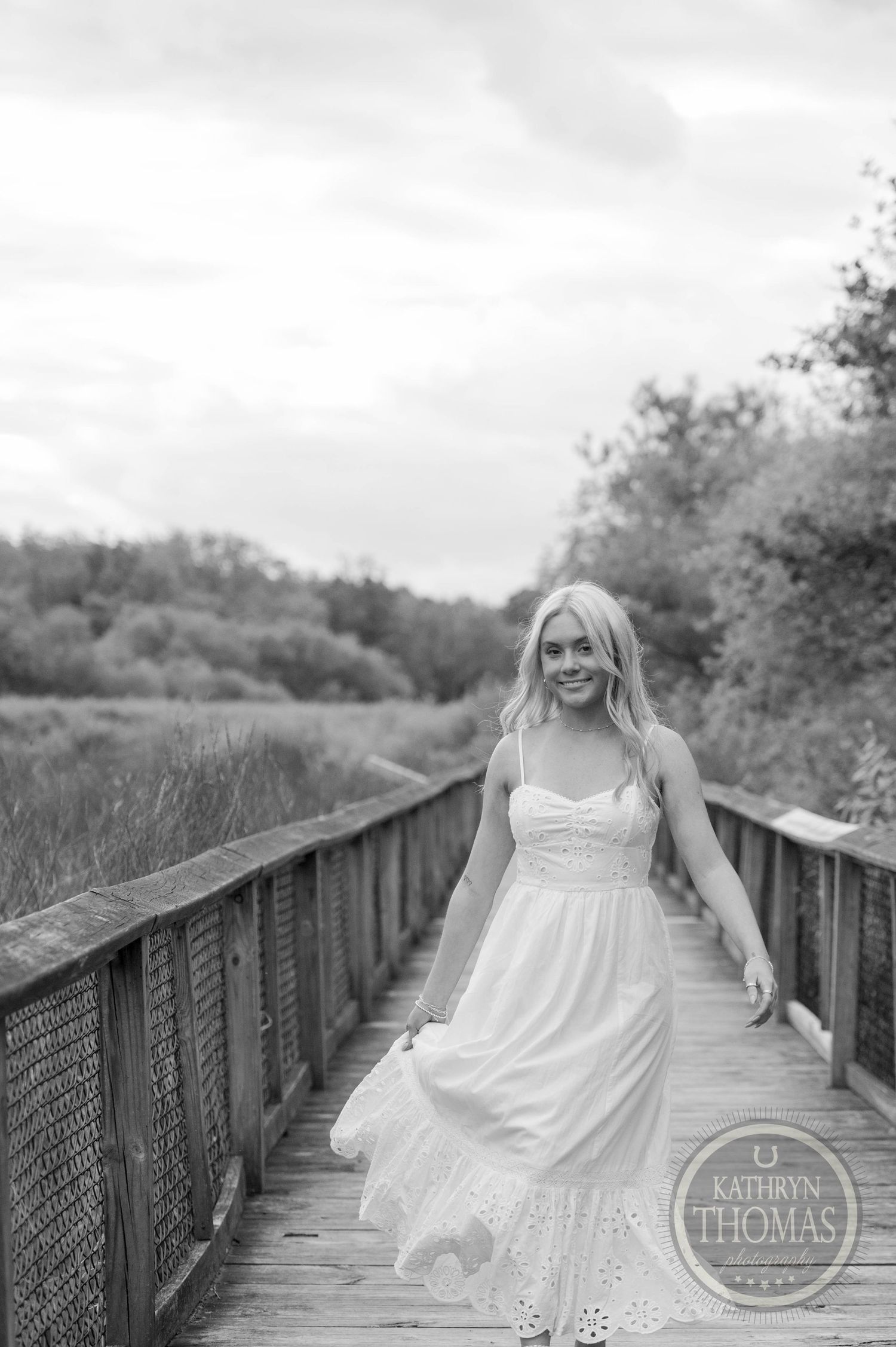 Person in white dress sitting on rustic wooden boardwalk surrounded by natural foliage in black and white photo.