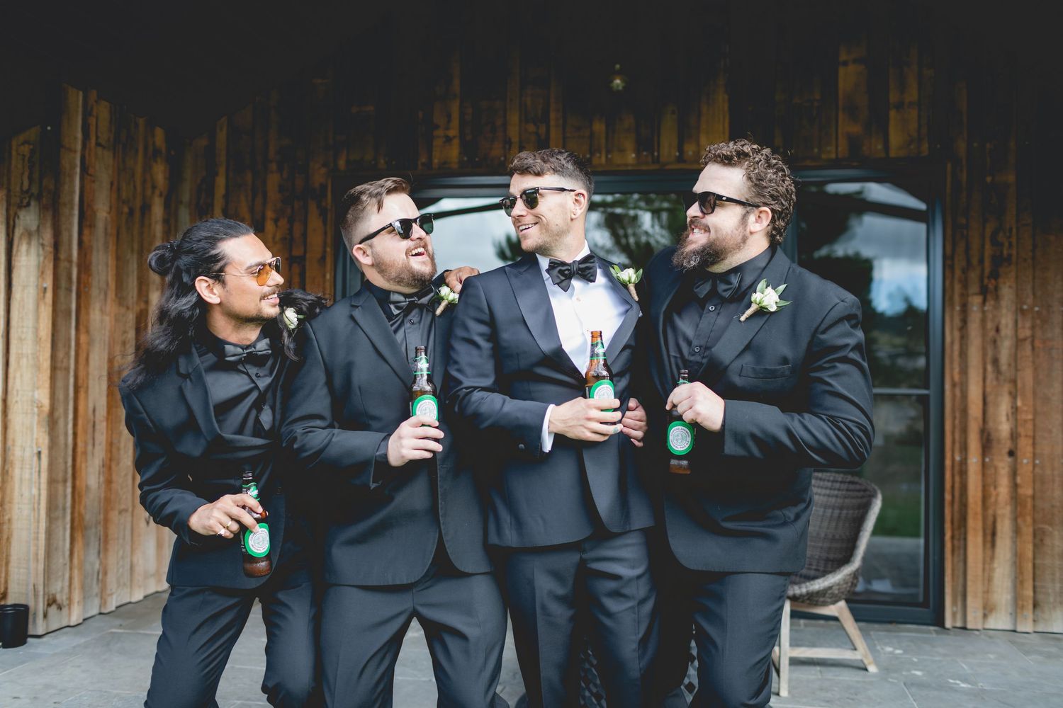 A group of four men in formal attire and sunglasses stand together laughing and holding drinks outside a wooden building.