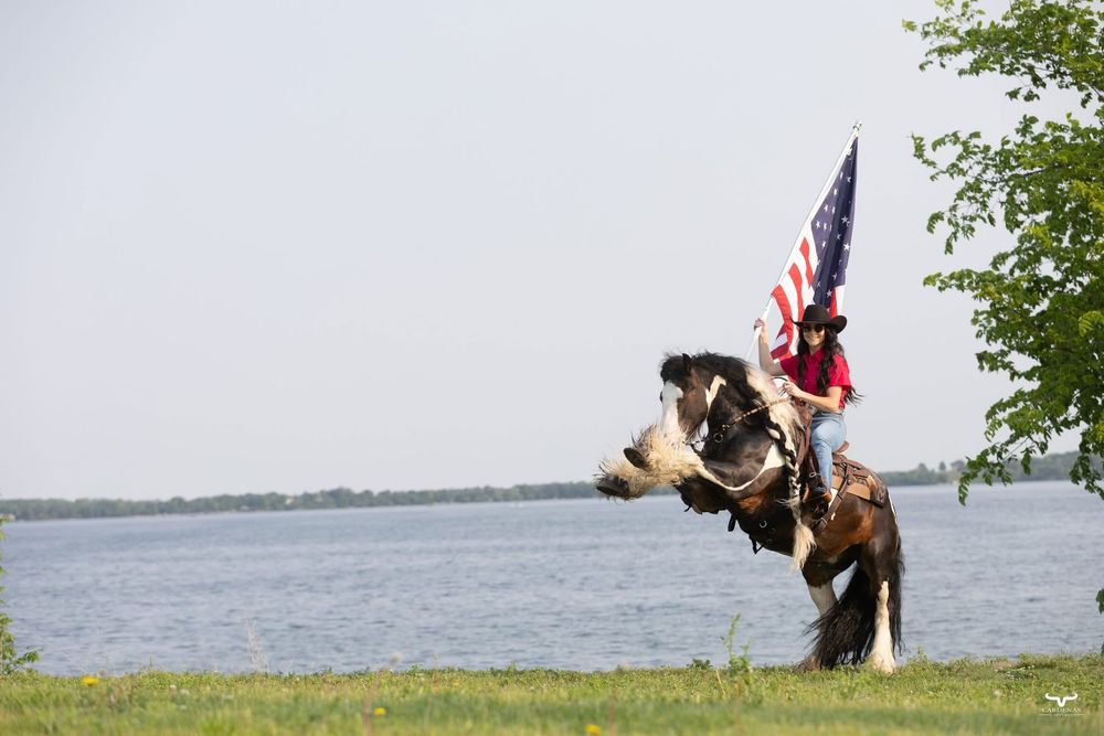 A horse and rider carrying an American flag stand by a lake. Pro Rodeo PRCA Rodeo Photographer Dakota Cardenas