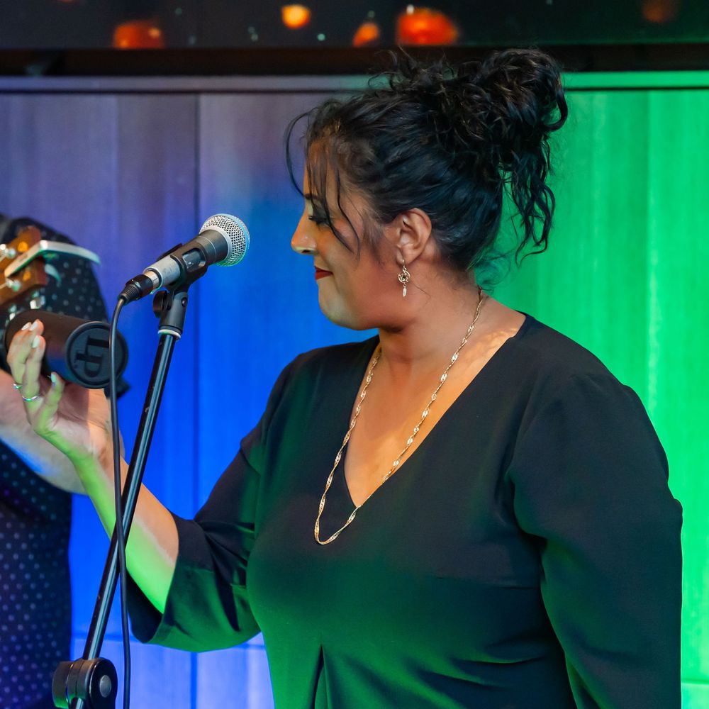 A performer in black attire sings passionately on stage with colorful blue and green lighting in the background.
