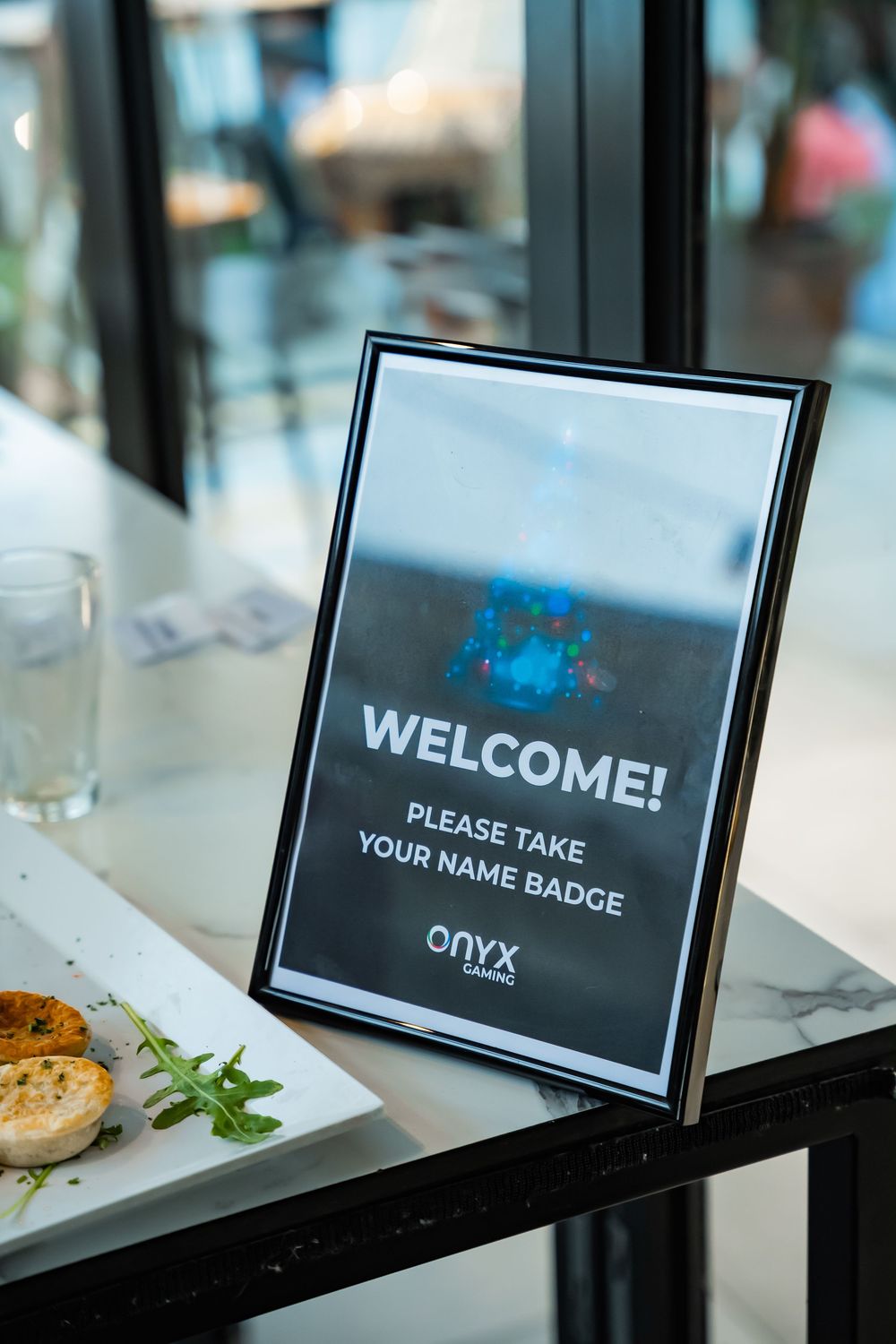 A welcome sign display on a table next to a plate of food asks guests to take their name badge at Onyx event.