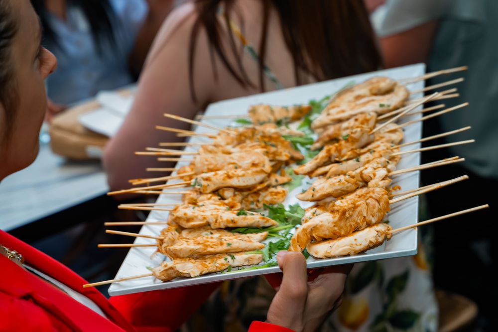 Server holding white platter of grilled chicken skewers garnished with fresh herbs at catered event.