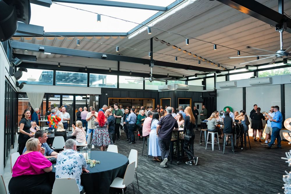 People gathered in a modern rooftop venue with tables and chairs enjoying a social event under a covered outdoor space.